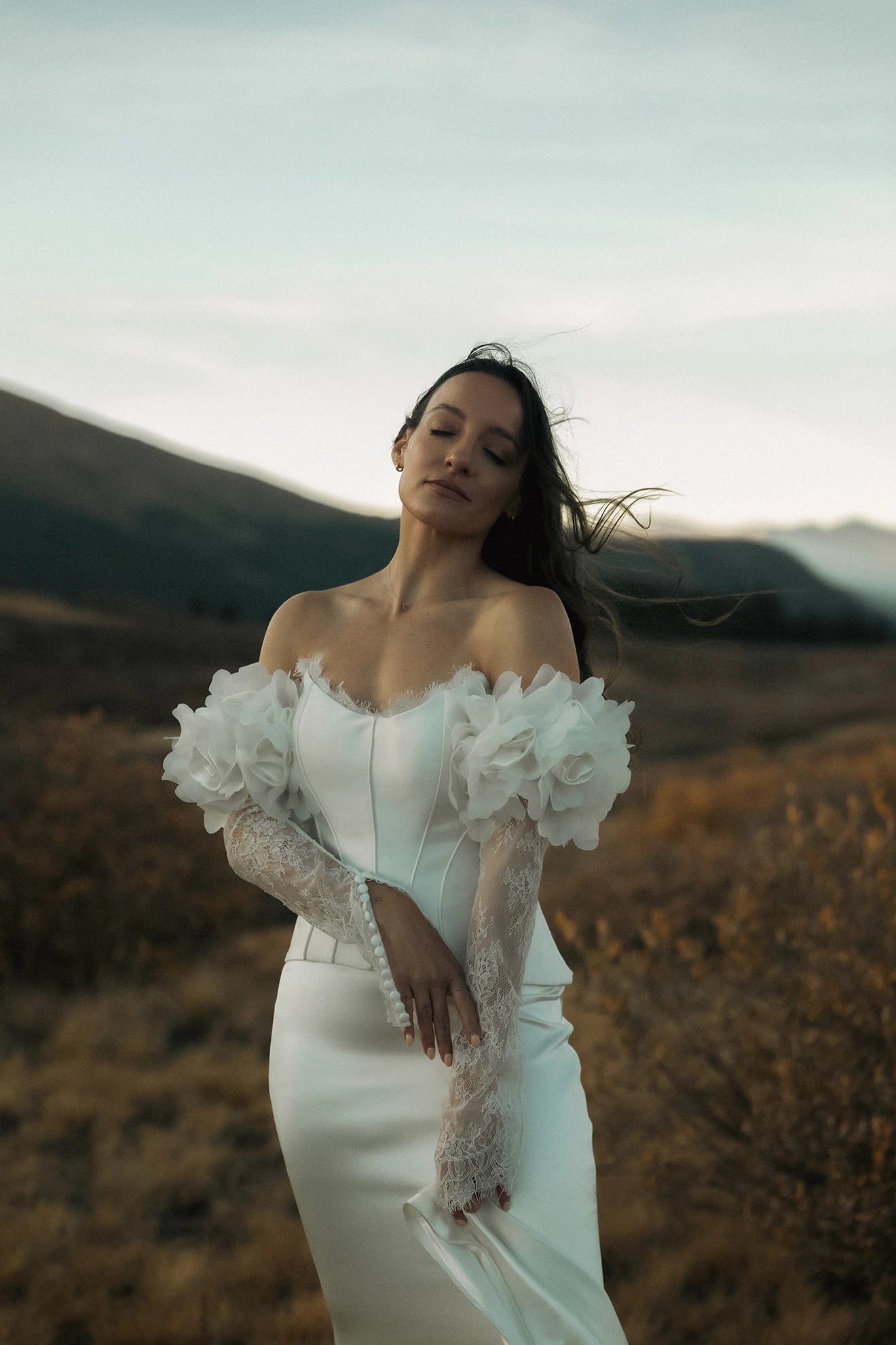 Woman in a white wedding dress with floral and lace sleeves standing in a field with mountains in the background