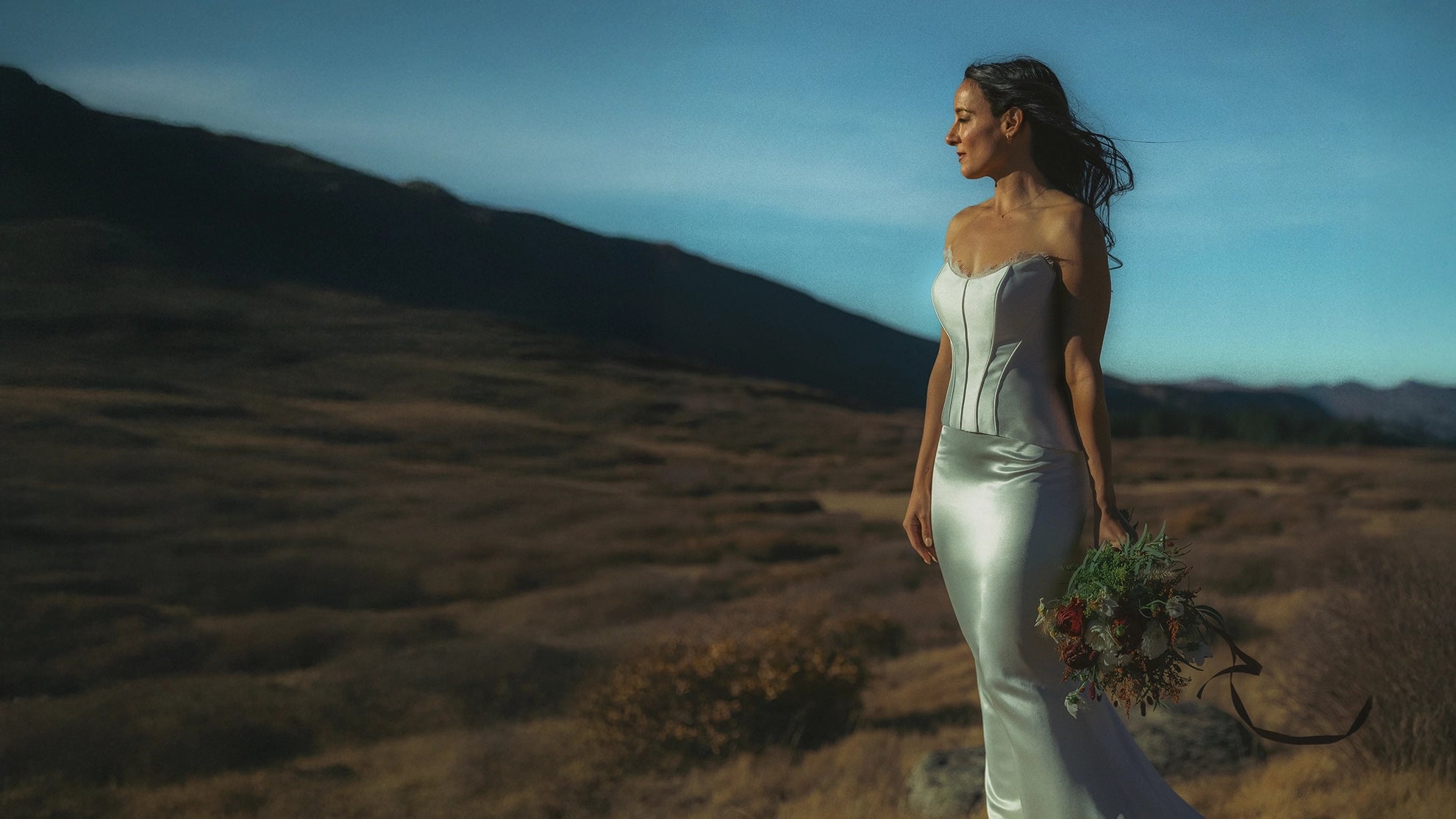 Woman in a white wedding dress holding flowers in a mountain landscape with mountains in the background