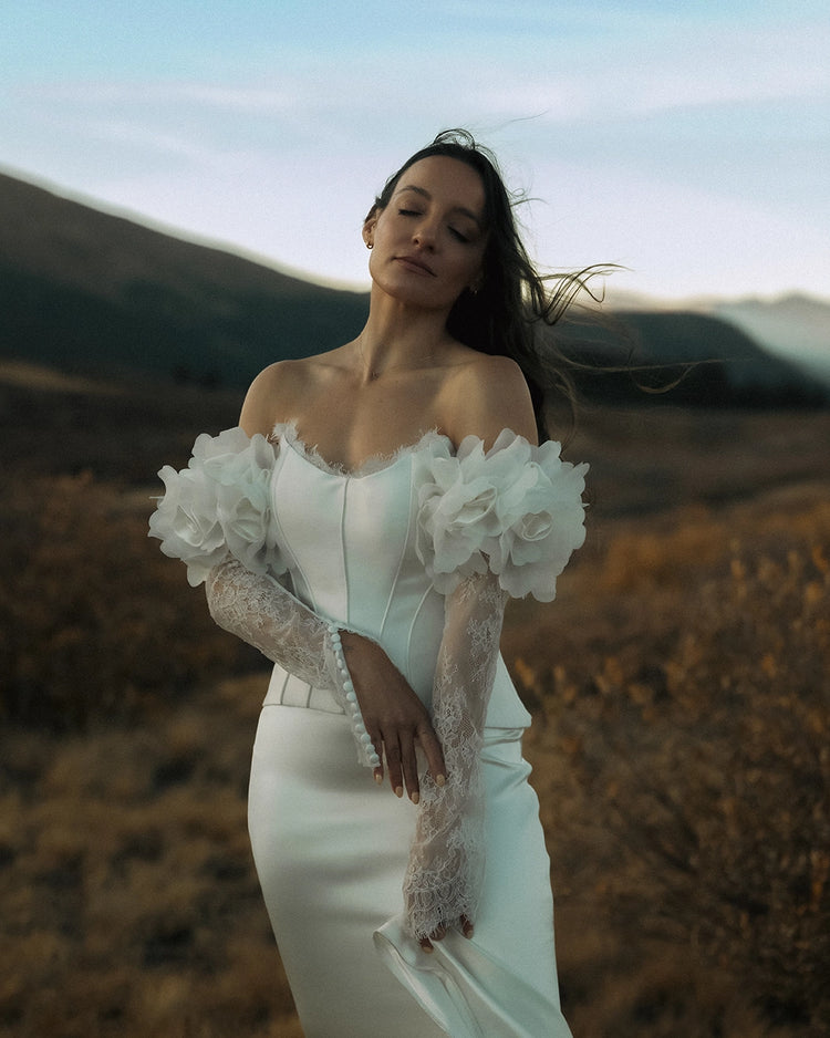 Woman in a white wedding dress with lace sleeves standing in a field with mountains in the background.
