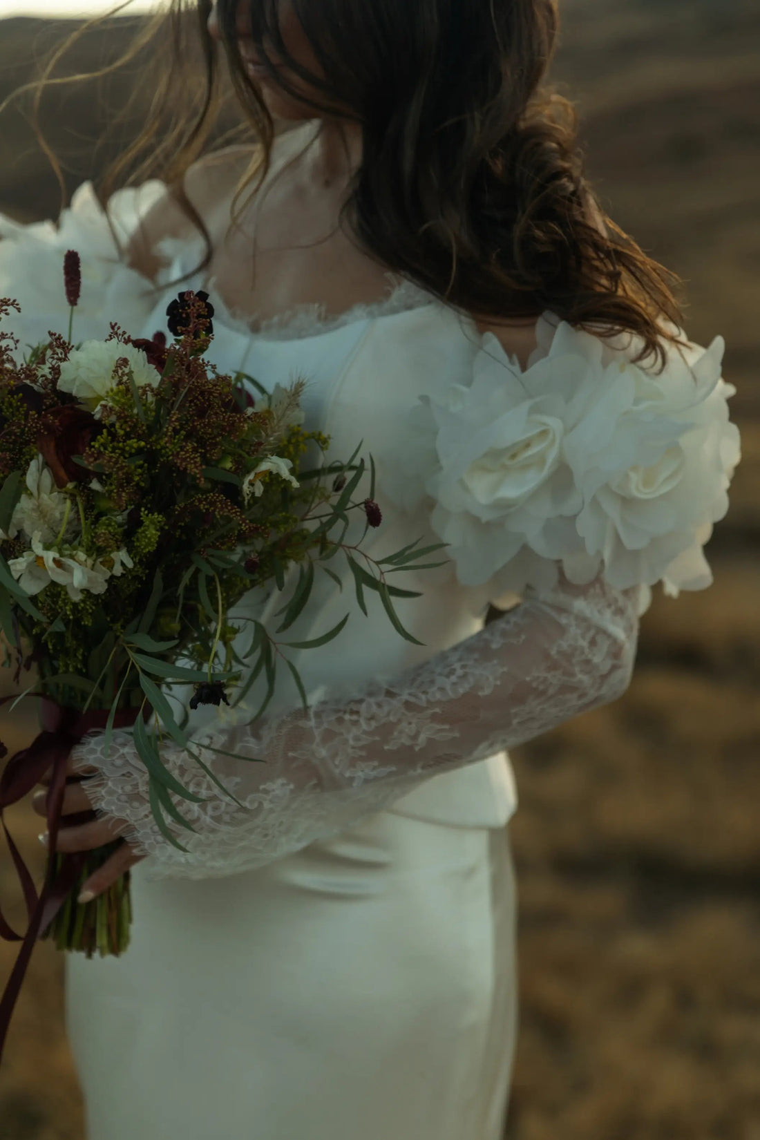 closeup of bride holding bouquet wearing a white wedding dress with floral and lace sleeves on top of blurred background