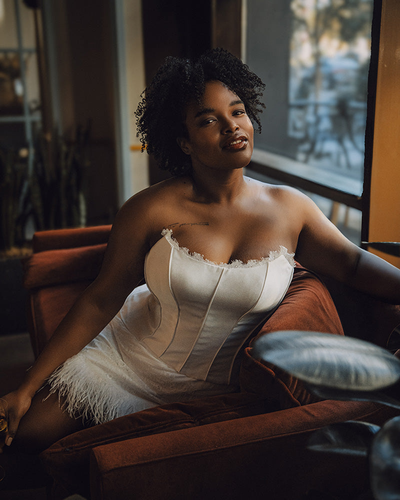Bride wearing a white corset sitting on a couch in a dimly lit room.